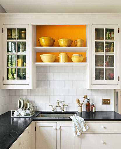 A mostly white-painted kitchen with black counter tops and orange accent behind shelving.