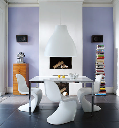 A modern dining room with a purple-painted accent wall, white fireplace, stacks of books, a dining table, and 3 modern dining chairs.