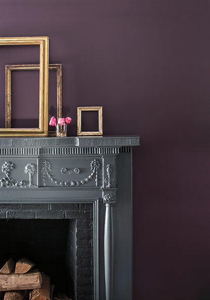 A gray fireplace in front of a purple-painted wall, with golden frames and pink flowers on the mantel.