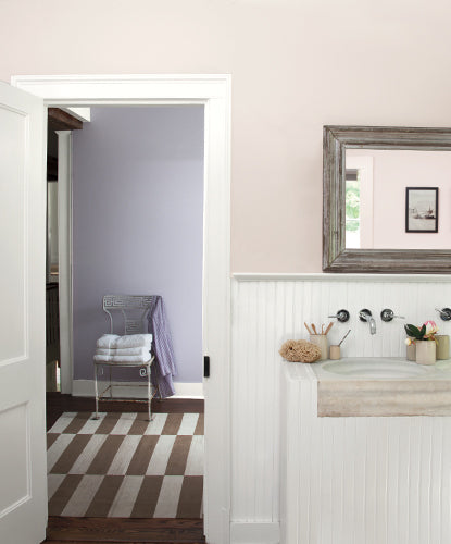 A bathroom with taupe walls tinged with light purple undertone; white wainscoting, cabinetry and trim; silver mirror; and white door open to a chair and rug in hallway.
