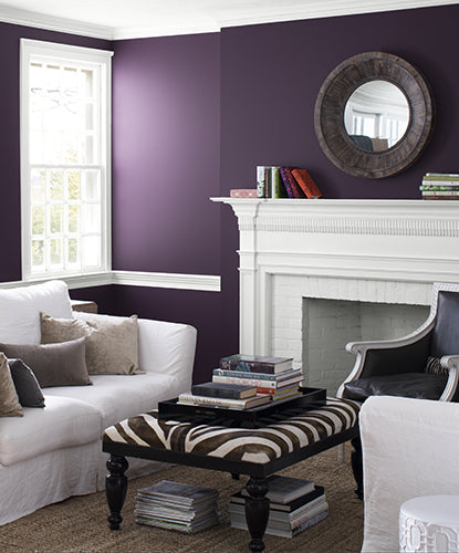 A purple-painted living room with white trim and fireplace, featuring white sofas, a blue armchair, and a zebra-print ottoman.