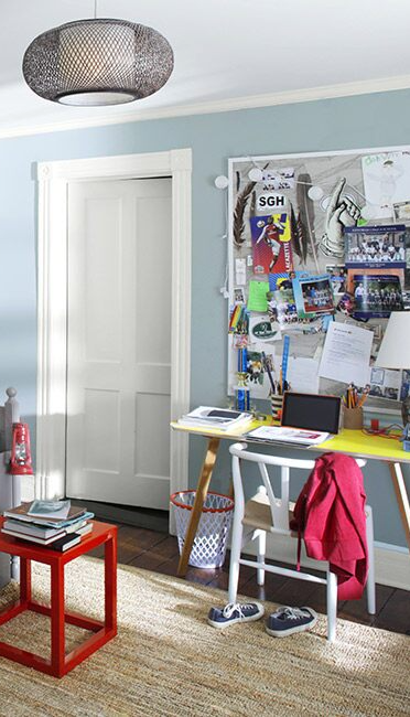 A gray-blue bedroom with white ceiling, cluttered bulletin board, bed with astronaut blanket, and red accent table.