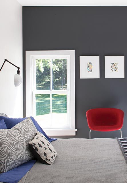A bedroom with a dark gray accent wall, red chair, gray bedding, and wall art.