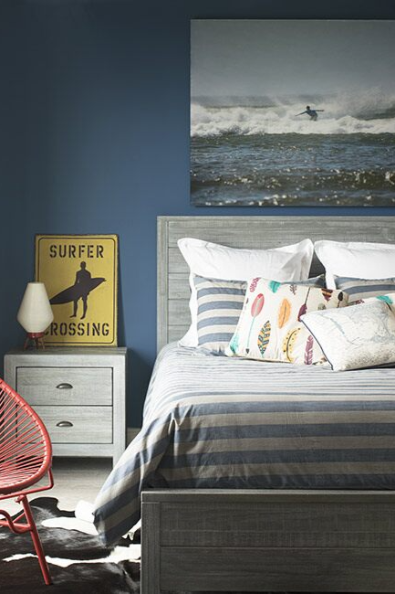 Bedroom with deep blue-painted walls, red accent chair, gray bedding, white accent table, and surfer-themed wall décor.