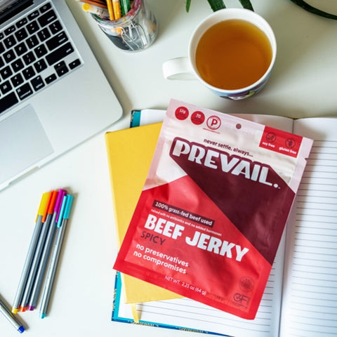 A work setup with a cup of tea and a pack of PREVAIL beef jerky