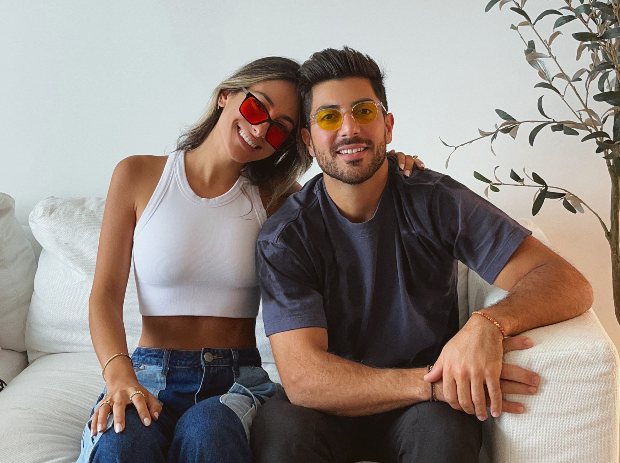 Two smiling people wearing colorful sunglasses sitting on a couch.