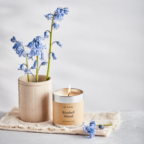 Lifestyle image of Bluebell Wood Scented Tin on table surrounded by vase of fresh bluebells