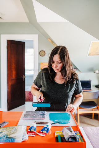 An image of a dark-haired woman rolling ink onto a printmaking block on an orange table.