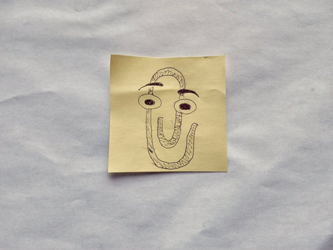 A yellow post-it note with a pen drawing of Clippy, an anthropomorphic paper clip with big eyes and floating eyebrows.