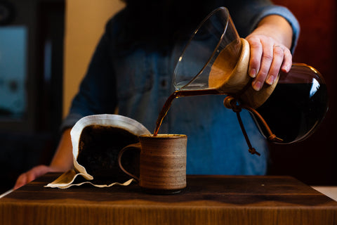 A person is pouring coffee from a Chemex into a ceramic mug. A cloth filter is in the background displaying the used coffee grinds.