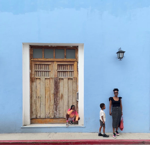 image shows a brown skinned woman and two children standing in front of a blue wall with a brown door.