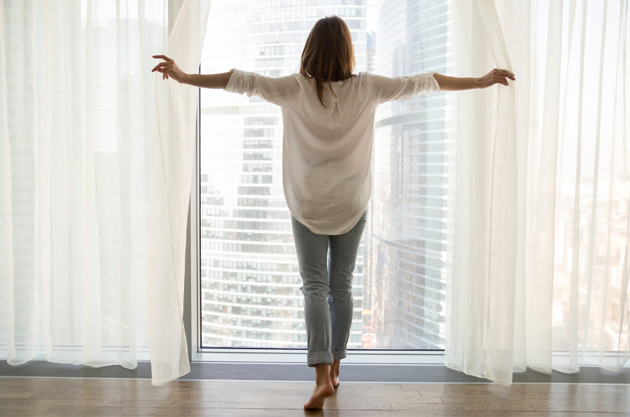 A woman opening window drapes to a cityscape view with information on how to properly hang curtains