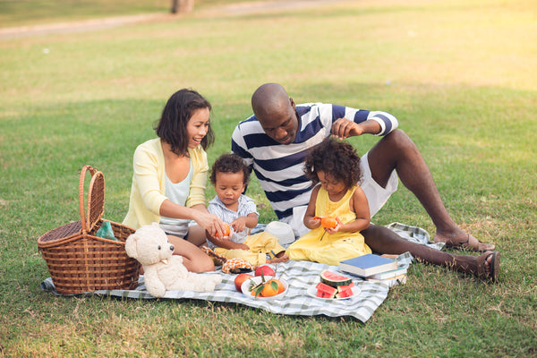 A family gathered on a picnic blanket with a picnic basket and various foods being enjoyed