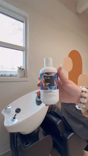 Person holding a bottle of IGK shampoo in a salon with a sink and colorful wall art.