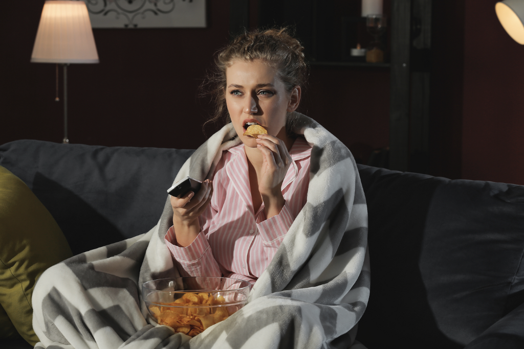 sleep procrastination: Woman sitting on a couch while watching TV and eating chips