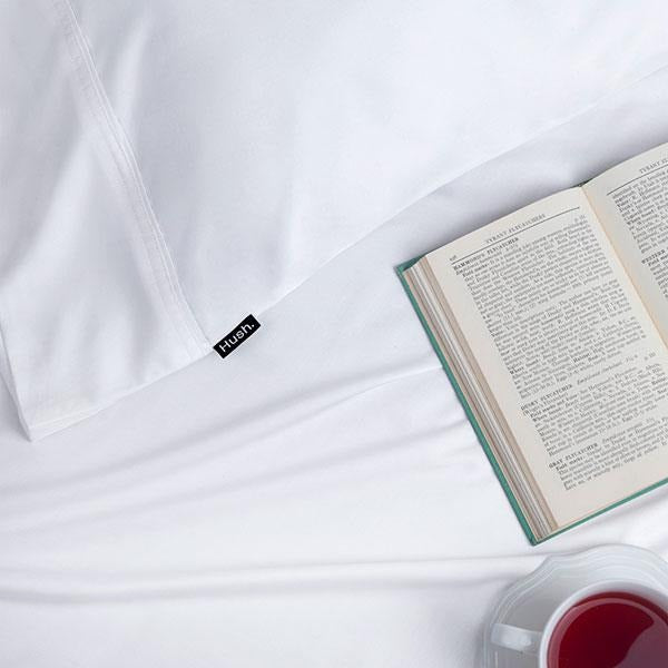An open book and a cup of tea sit on a bed made with Hush Iced 2.0 Cooling sheets. A Hush tag is visible on the pillowcase.