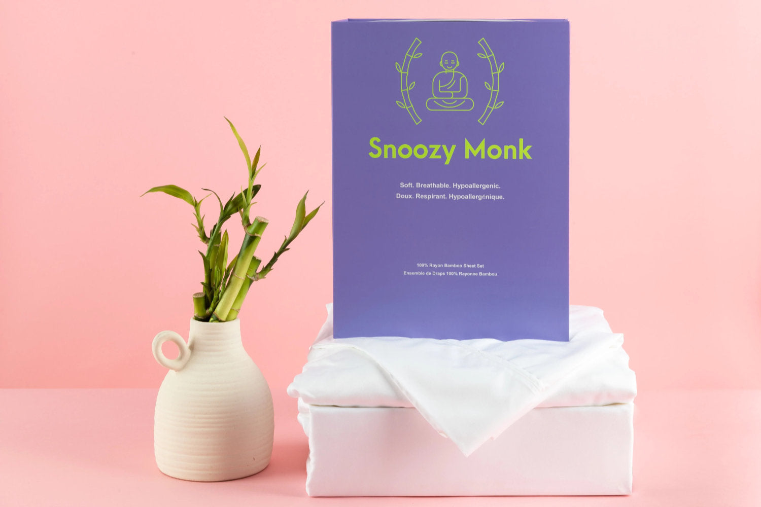 Snoozy Monk Bamboo sheets folded in a square. A bamboo plant sits next to the sheets. A Snoozy Monk informational card sits on top of the sheets. It reads “Snoozy Monk ⁄ Soft. Breathable. Hypoallergenic. ⁄ Doux. Respirant. Hypoallergénique. ⁄ 100% Rayon Bamboo Sheet Set ⁄ Ensemble de Draps 100% Rayonne Bambou”