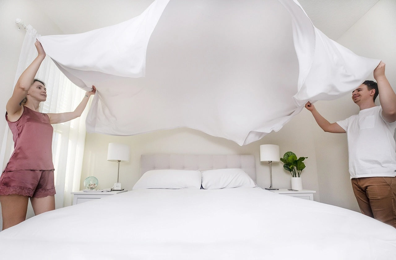 A man and a woman stand on opposite sides of a bed, holding a bamboo sheet aloft as they bring it down onto the bed.