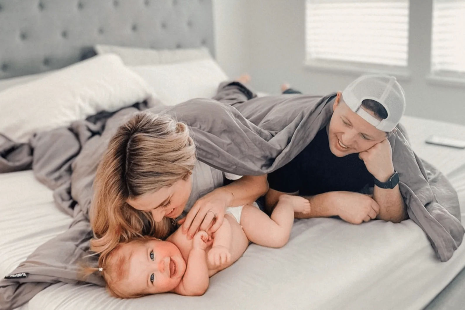 Mom and dad with their baby smiling happily in bed while sharing a grey Hush Iced Cooling Weighted Blanket.