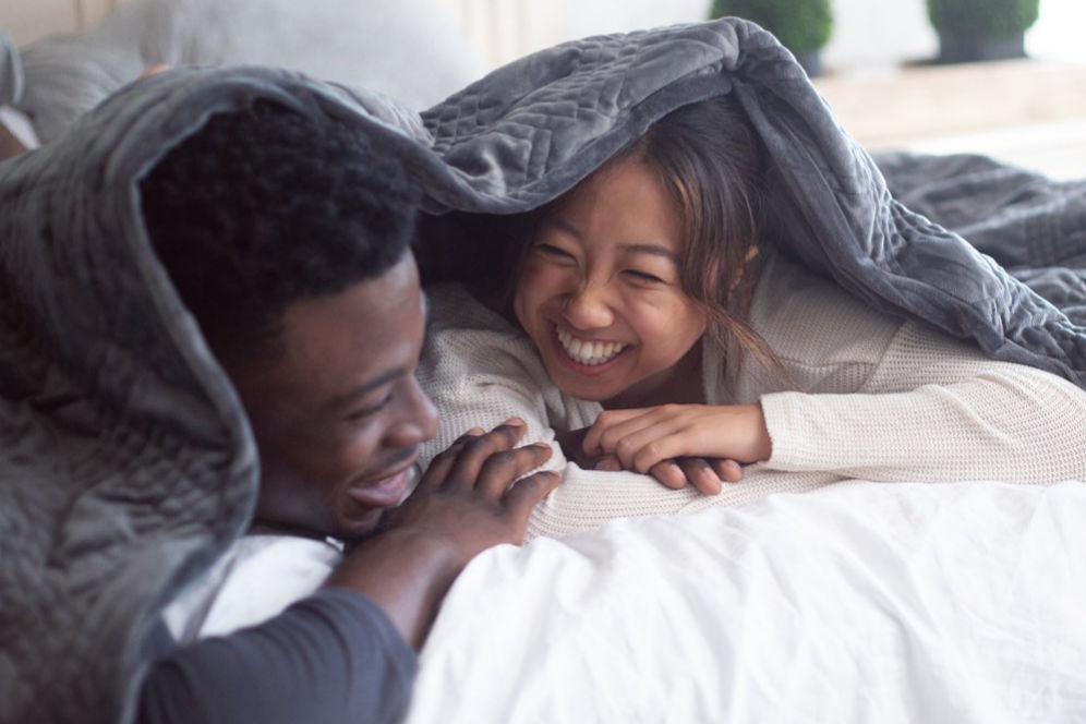 A black couple lying on their stomach in bed and smiling at each other while under a Hush Classic Weighted Blanket.