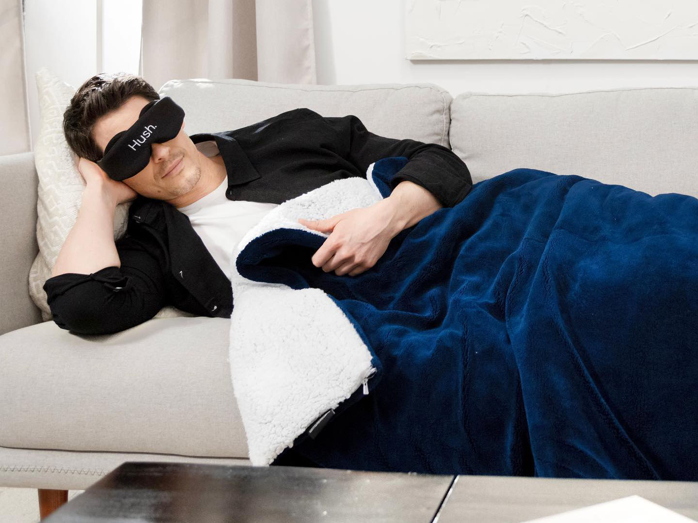 A man sleeping under a weighted blanket wearing a Hush-branded blindfold.