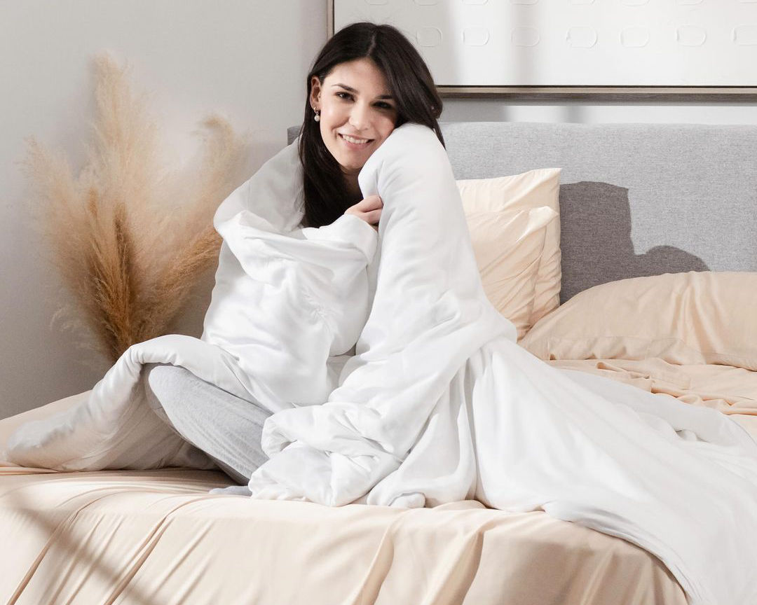 A woman sitting in bed smiling and snuggling a Hush weighted blanket.