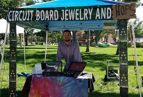 Drew smiling at his booth at an art fair on a sunny day at the park. A sign hangs above his head that reads "circuit Board Jewelry and Hops" He is surrounded by TechWears Circuit Board Products.
