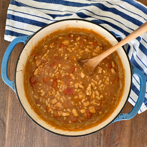 Chicken chili in a blue Dutch oven with a wooden spatula in it, on top of a wooden table with a blue-stripped towel nearby