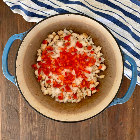 Blue Dutch oven pot on a wooden table filled with brown chicken and topped with chopped onions and red bell pepper. There is blue stripped towel in the corner.