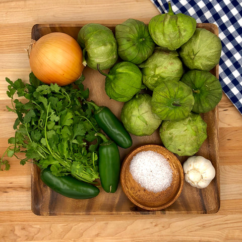 ingredients to make salsa verde on a wooden plate. includes: tomatillos, onion, cilantro, jalapeno, garlic, salt