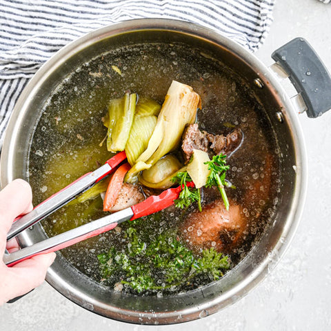 All of the ingredients in a large pot with water, featuring hands stirring the soup with red tongs. 