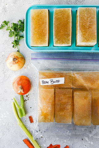 Close up shot of 1-cup Souper Cubes tray at the top with frozen broth with 1-cup portioned broth cubes in a large Ziplock bag with a piece of masking tape labeled “bone broth”. 