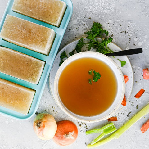 1-cup Souper Cubes trays with frozen oxtail bone broth placed in the left hand corner with a bowl of thawed broth in a white bowl.