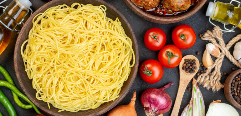 A bowl of pasta next to tomatoes, red onions, and related ingredients and utensils.