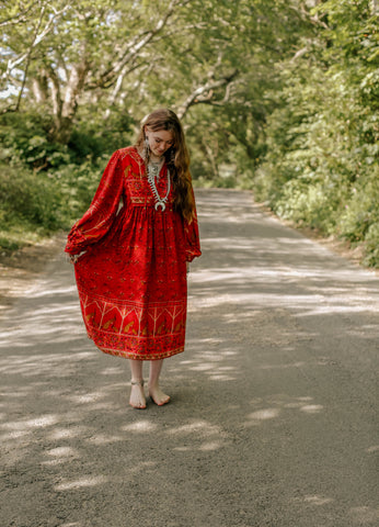 Photograph of a model in a rural setting wearing a 1970s style bohemian red midi dress with balloon sleeves.