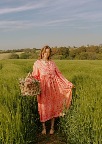 Photograph of a model in a field wearing a long bohemian style pink midi dress with big balloon sleeves holding a straw basket.