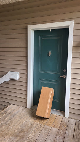 A blue door with white trim is shown next to a flight of outdoor, wooden plank stairs. There is a plain brown box leaning up against the door that is about half the height of the door. It is plain and brown with no distinguishable markings on it.