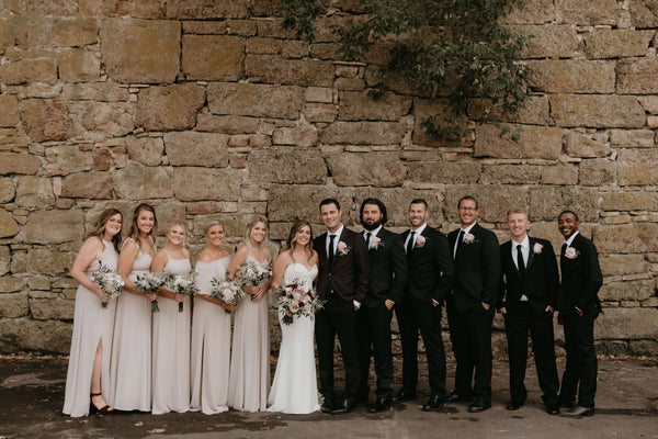 Bridal Party lined up in front of a beautiful stone wall. Groomsmen are wearing black suits. 