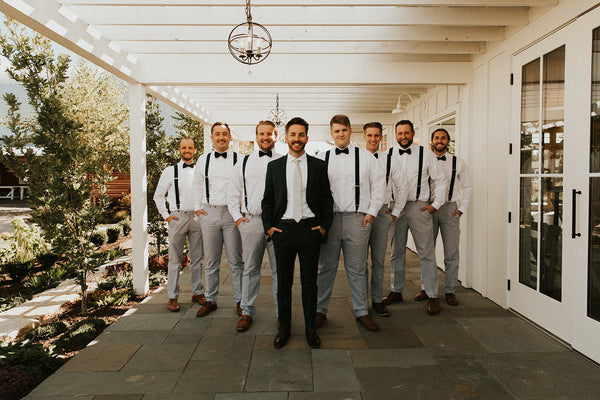 Groom is wearing a black suit and white tie. Groomsmen are wearing grey dress pants and black bowties. 