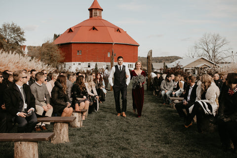 Ceremony is set outside in a woodsy area. The pews are made of wood and have tree stumps as support. Bridesmaid and Groomsman walking down the aisle. 