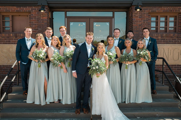 Bridal Party posing together on stairs in front of brick building. Bride is wearing 'Rebecca Ingram Raelynn' Dress from the Wedding Shoppe.