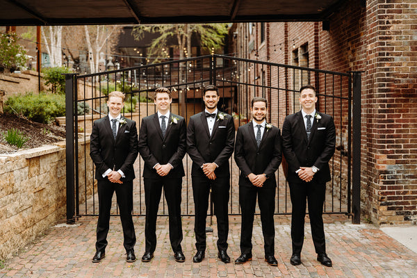 Groomsmen in black tuxedos lined up on a brick pathway outside of a brick building. 