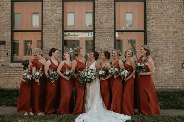 Bride and Bridesmaids gathered outside a brick building, smiling and laughing with one another. 
