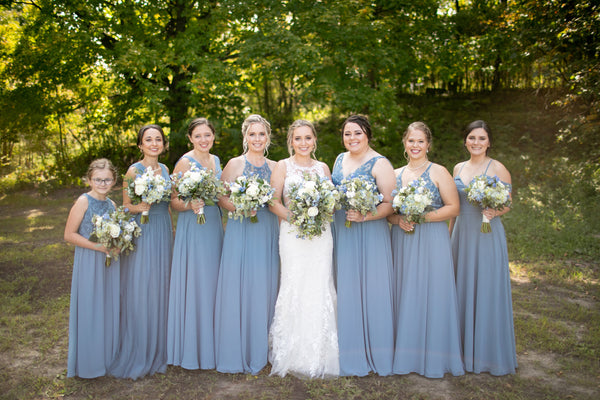 Bride and Bridesmaids wearing long flowy light blue dresses gathered outside around lots of greenery.