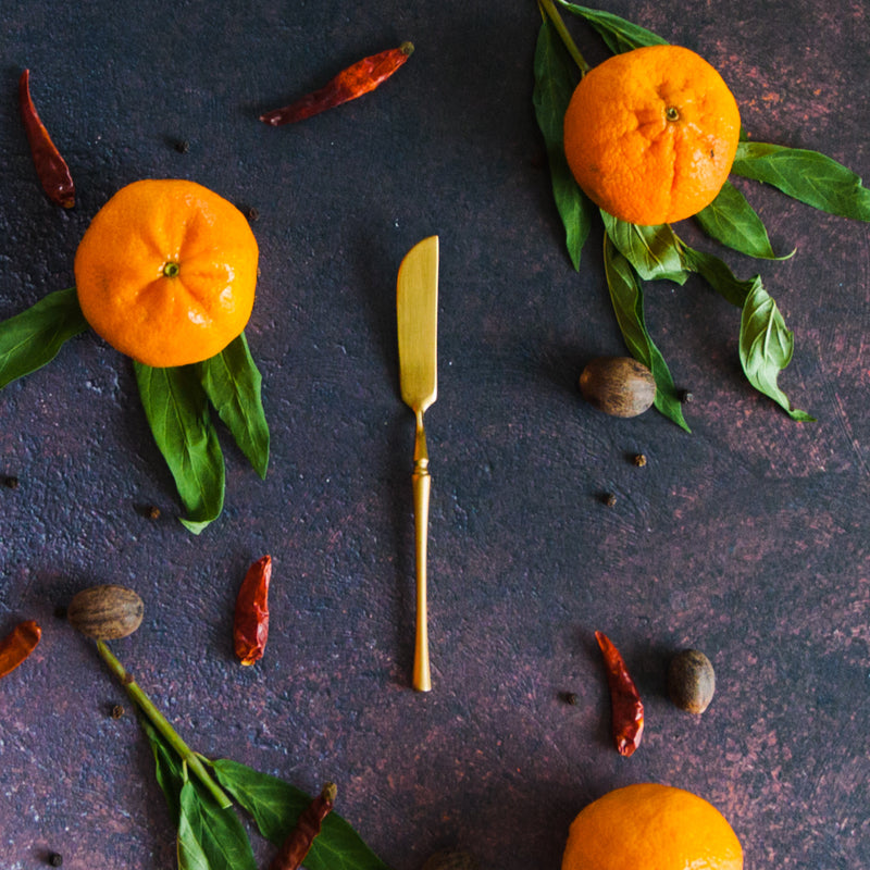 Gold Cheese knife on a rusty black surface with clementines, green leaves and red chilis