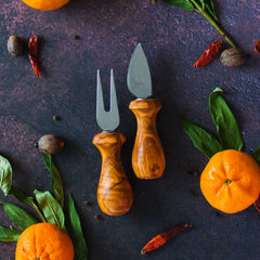 Pair of olive wood cheese knives on a dark background with Mandarin oranges, green leaves and red chilis