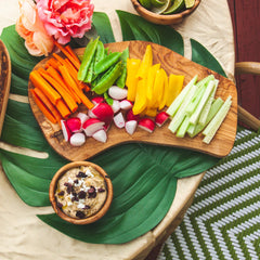 Olive Wood Cheese board with a rainbow of veggies arranged on it, on a green leaf placemat alongside a bowl of hummus