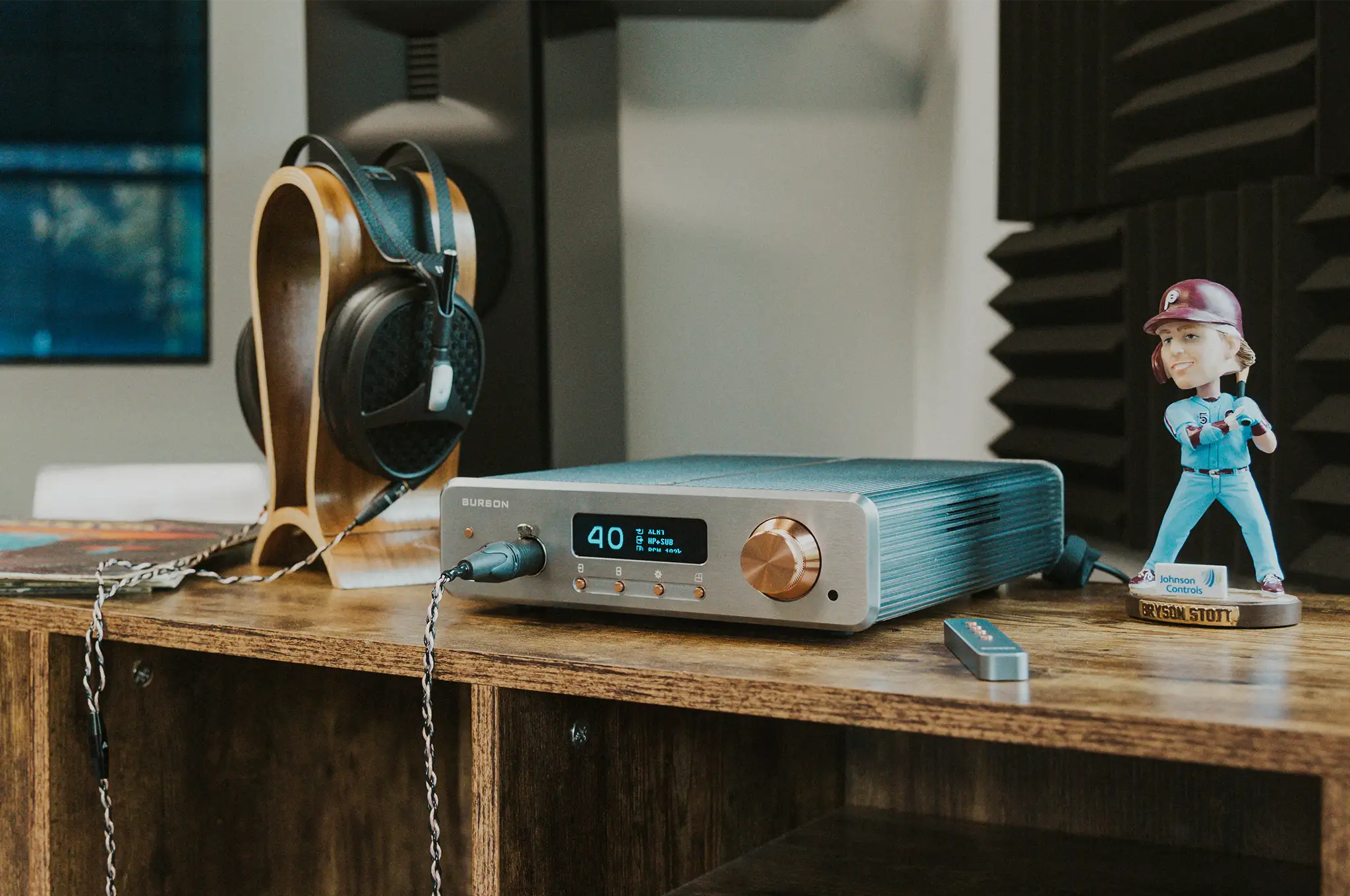 Burson Audio Conductor GT4 amp sitting right 3 quarter angle on wooden table with meze empyrean headphone on stand, bobble head, remote and stack of vinyl records