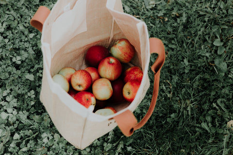 Fabric grocery bag filled with apples on grass.