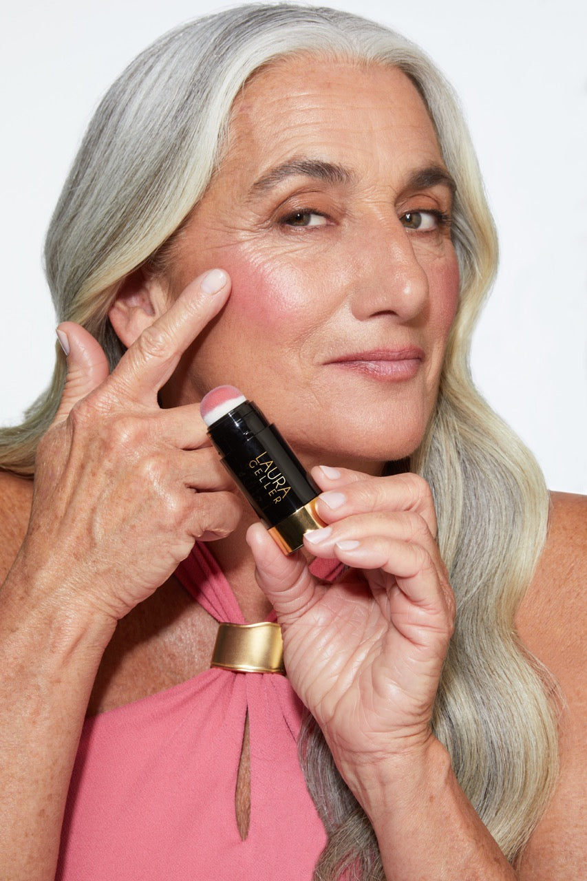 Elderly woman with grey hair holding a makeup stick near her cheek.